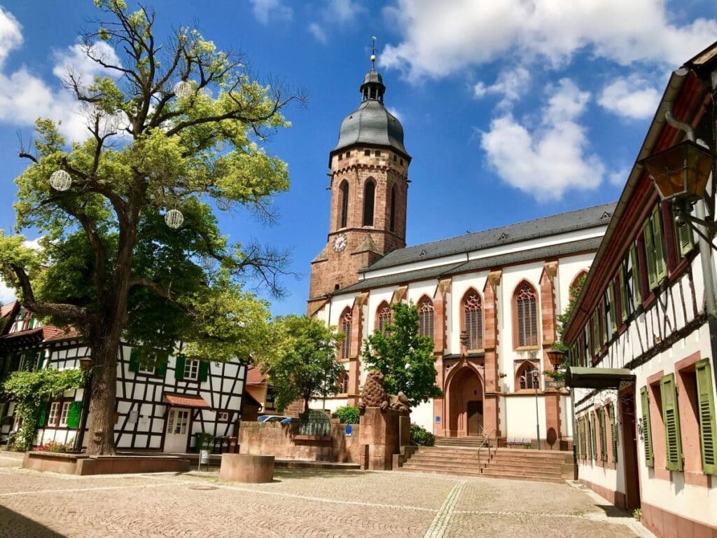 Im Herzen von Kandel liegt ein malerischer Dorfplatz mit einer bezaubernden Kirche, Fachwerkhäusern und einem Baum, der sich anmutig unter einem blauen, wolkenverhangenen Himmel streckt.