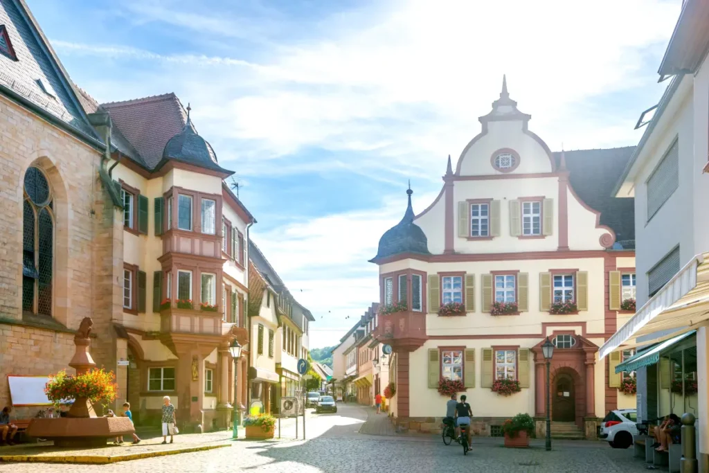 Eine sonnige Straße mit Kopfsteinpflaster, gesäumt von charmanten, historischen europäischen Gebäuden und Menschen, die spazieren gehen oder Rad fahren.