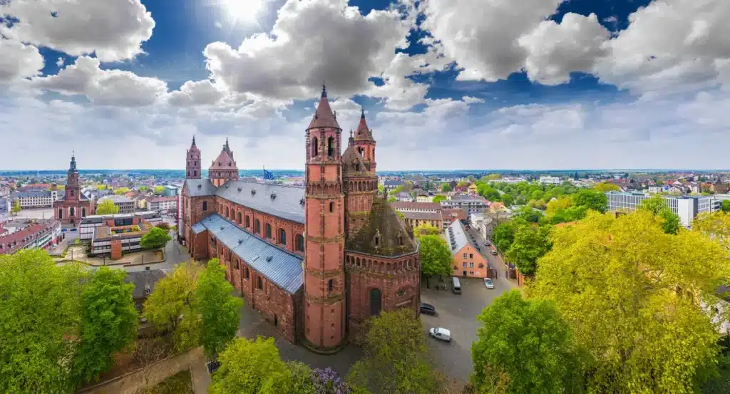 Luftaufnahme einer großen, historischen Kathedrale, umgeben von Bäumen und städtischen Gebäuden unter einem teilweise bewölkten Himmel.