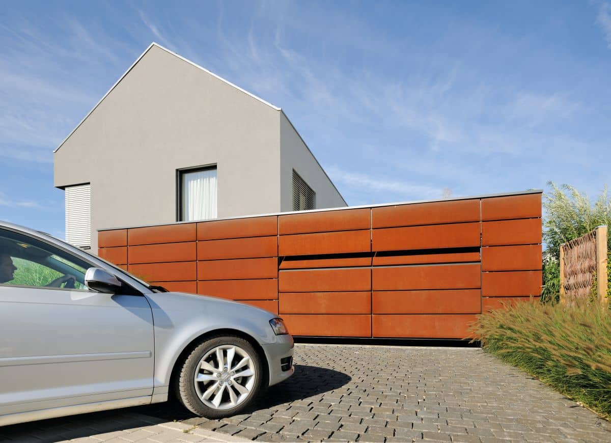 Modernes Hausäußeres mit rostfarbenem Sektionaltor, graues Auto parkt auf der gepflasterten Auffahrt unter einem klaren blauen Himmel.