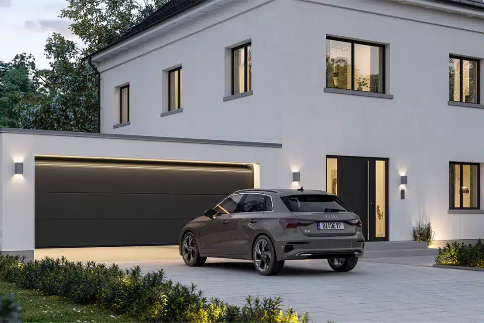 Ein modernes graues Auto, das vor einem weißen Haus geparkt ist, akzentuiert durch ein elegantes schwarzes Garagen-Sektionaltor, fängt die heitere Eleganz der Abenddämmerung ein.