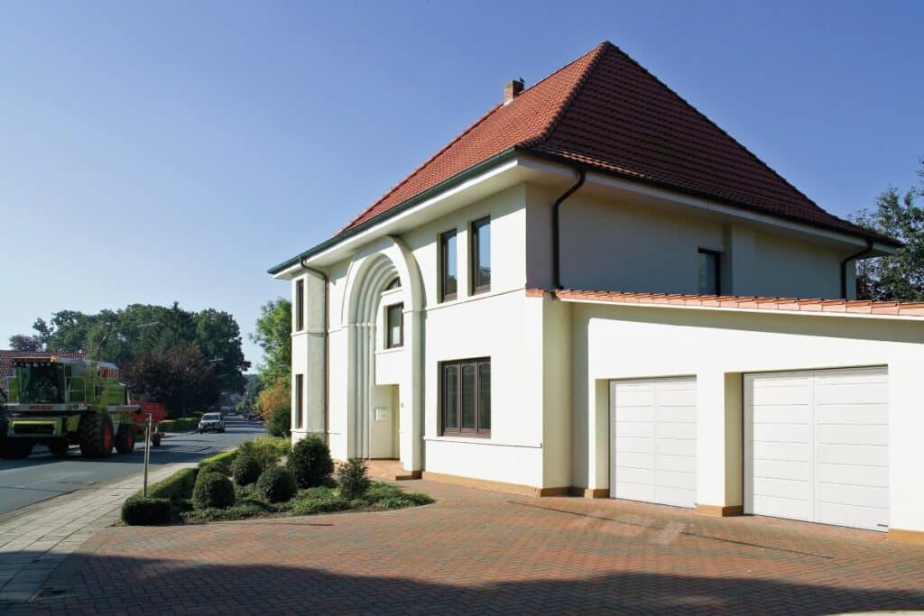 Modernes zweistöckiges Haus mit rotem Dach, Garage und einem Traktor auf der Straße im Hintergrund an einem sonnigen Tag.