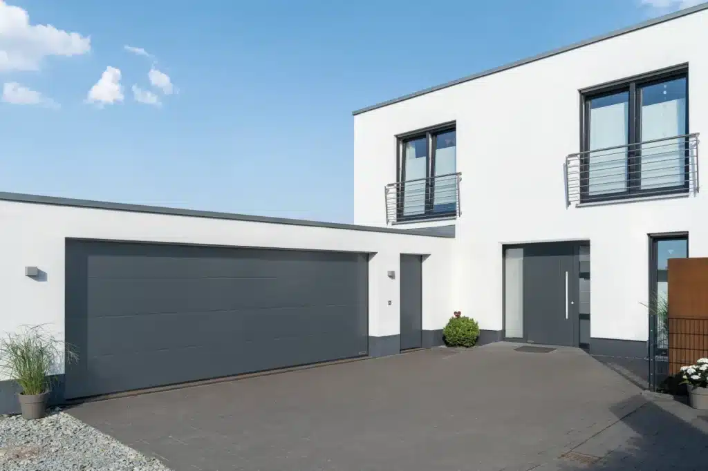 Modernes Haus mit einem großen grauen Garagentor, weißen Außenwänden und zwei Fenstern im Obergeschoss.