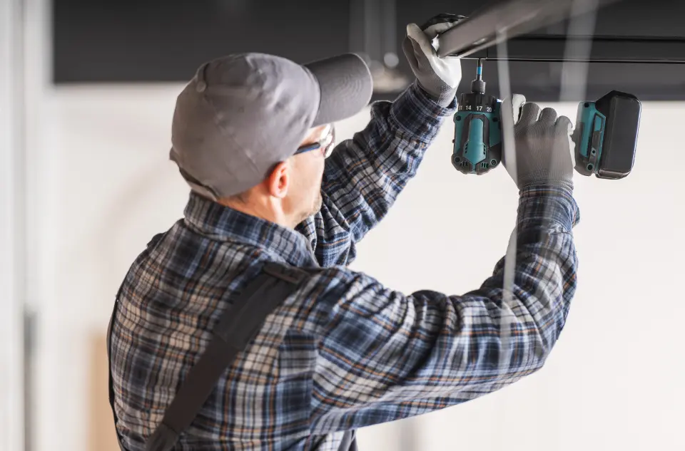 Arbeiter in kariertem Hemd und Mütze bedient mit einer Bohrmaschine den Mechanismus eines Garagentors im Innenbereich.