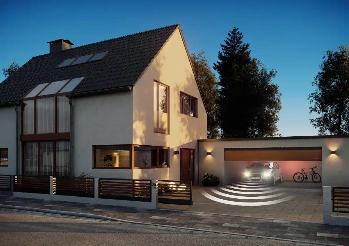 Modernes Haus mit beleuchteter Garage, einem darin geparkten Auto und Fahrrädern in der Nähe. Abendstimmung mit Bäumen im Hintergrund.