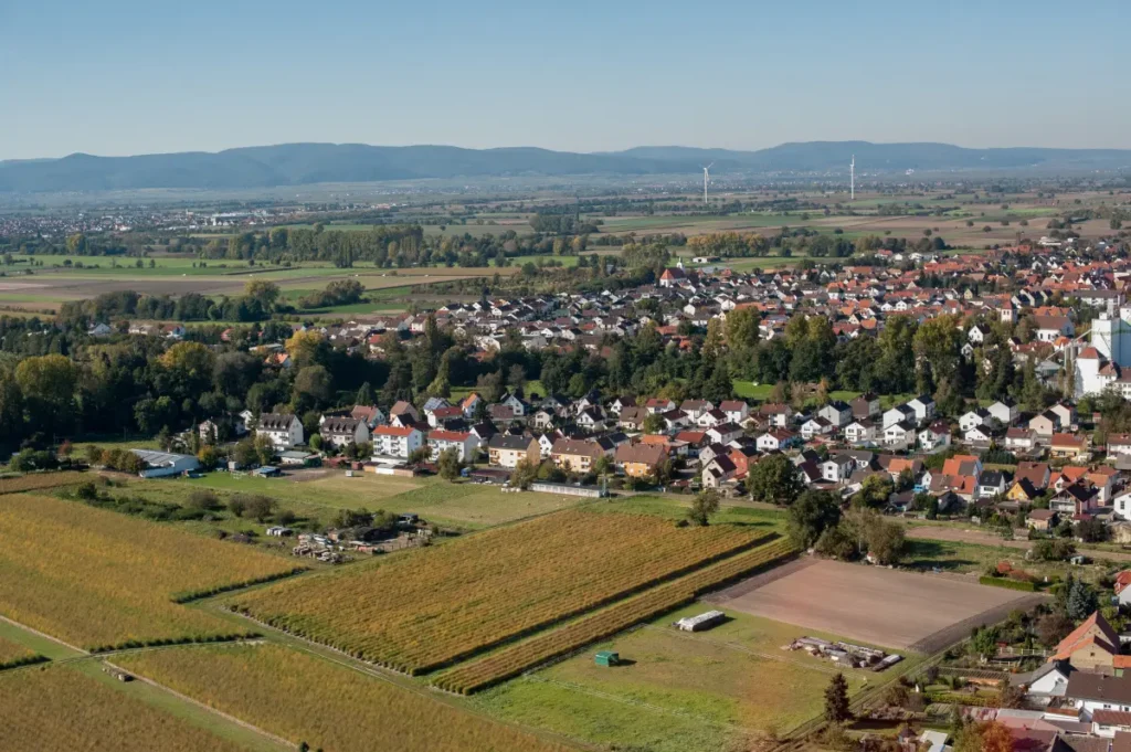 Luftaufnahme einer kleinen Stadt mit Häusern, Feldern und entfernten Bergen unter einem klaren blauen Himmel.