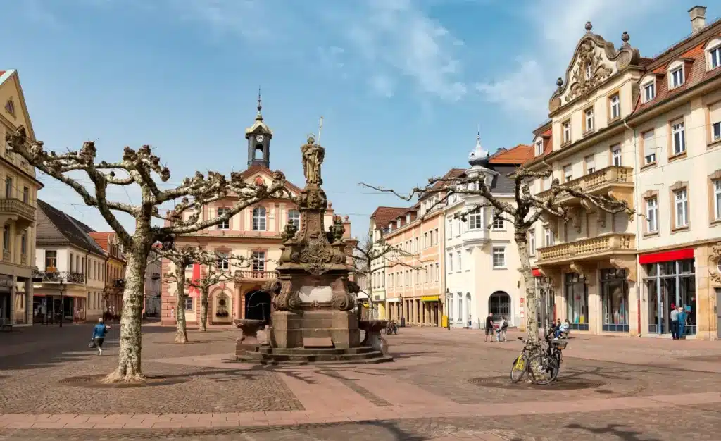 Ein Stadtplatz mit einer Statue, beschnittenen Bäumen, historischen Gebäuden und ein paar Menschen, die spazieren gehen oder Rad fahren.