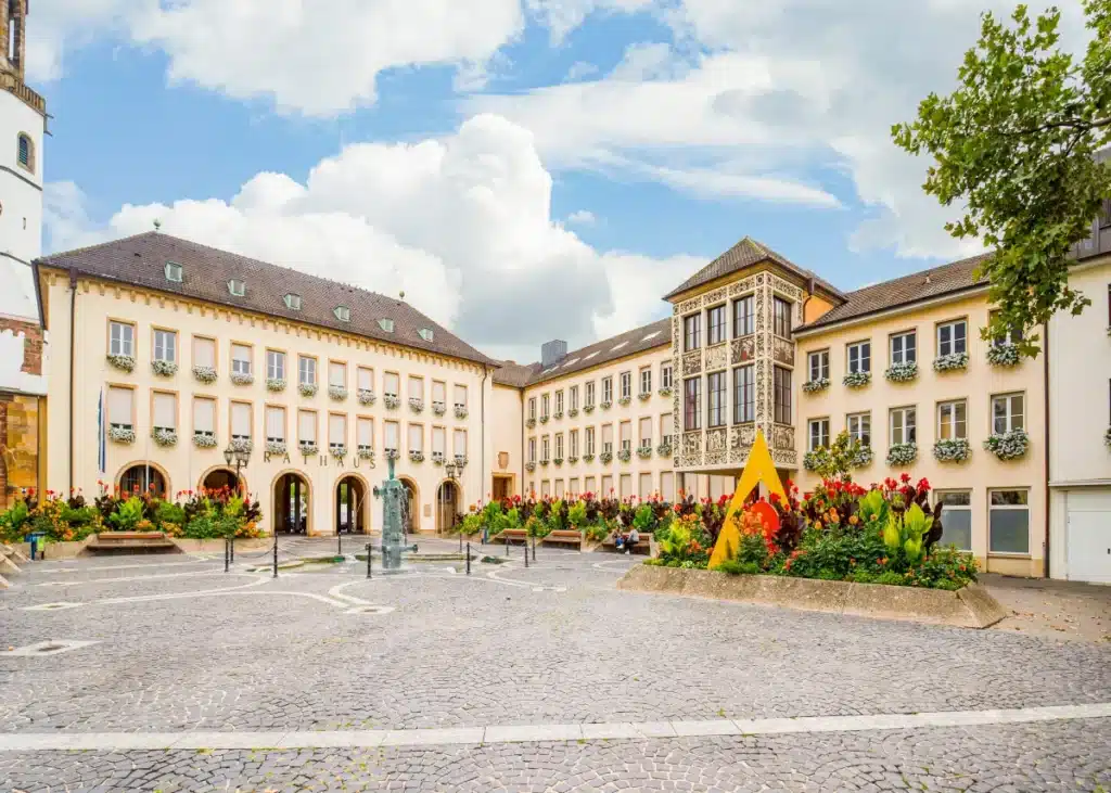 Ein kopfsteingepflasterter Platz mit Blumenbeeten, einer gelben Skulptur und einem historischen Gebäude mit vielen Fenstern und Balkonen.