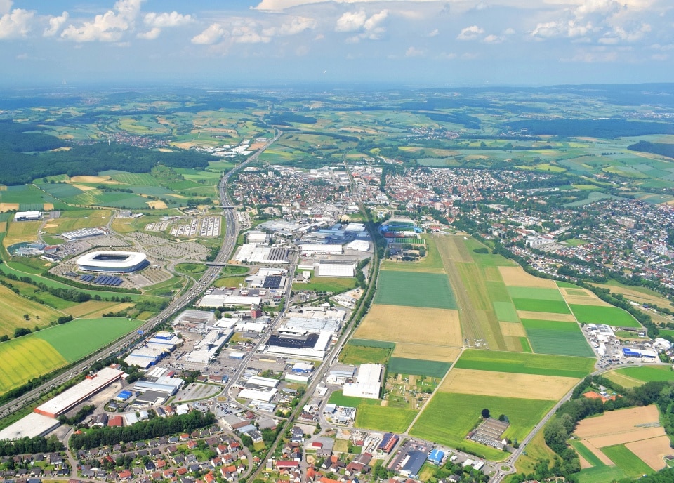 Luftaufnahme einer Stadt mit einem Stadion, Feldern, Industriegebäuden und Wohngebieten unter blauem Himmel.