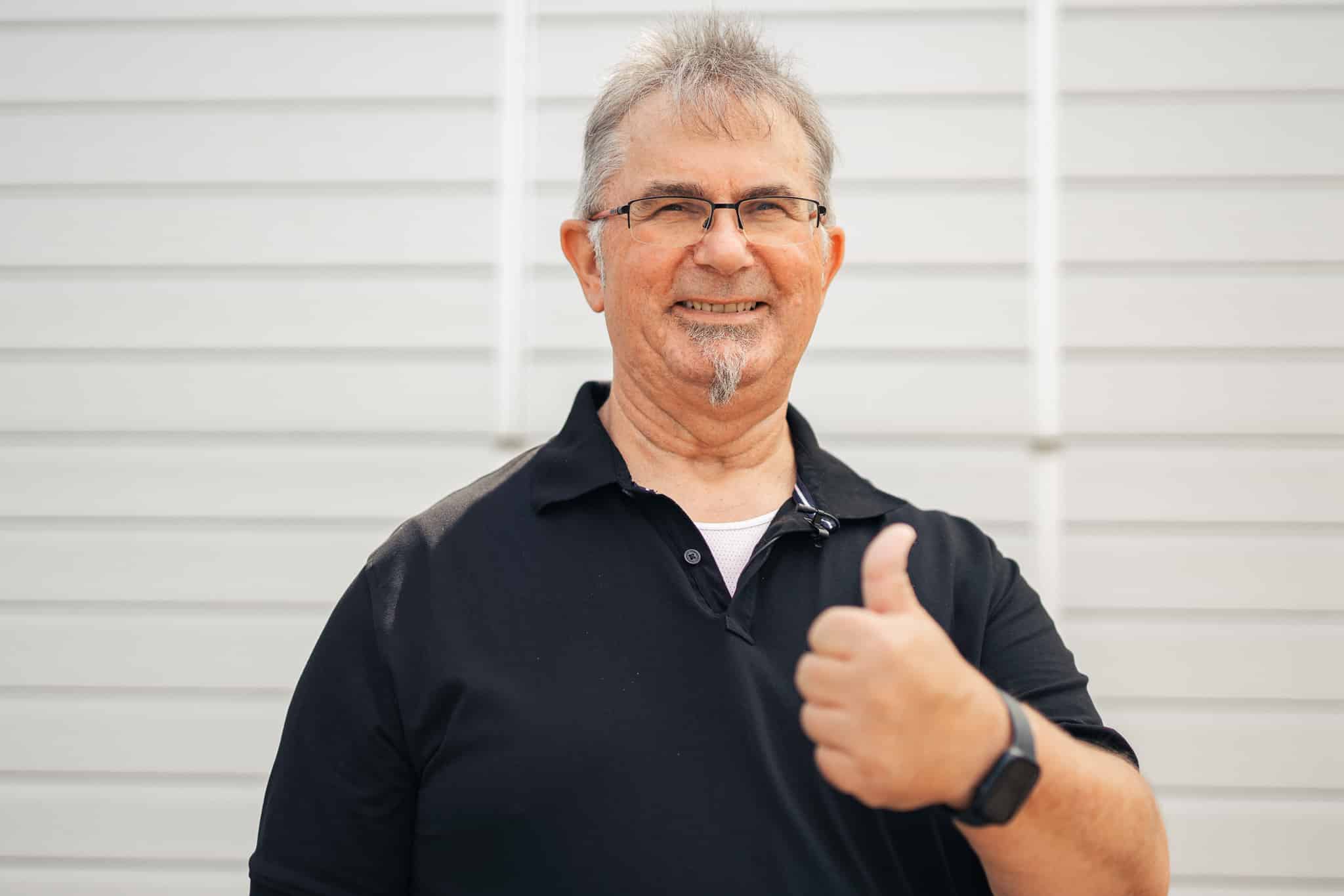 Lächelnder älterer Mann mit Brille und schwarzem Hemd, der vor einer hellen Wand den Daumen nach oben streckt.