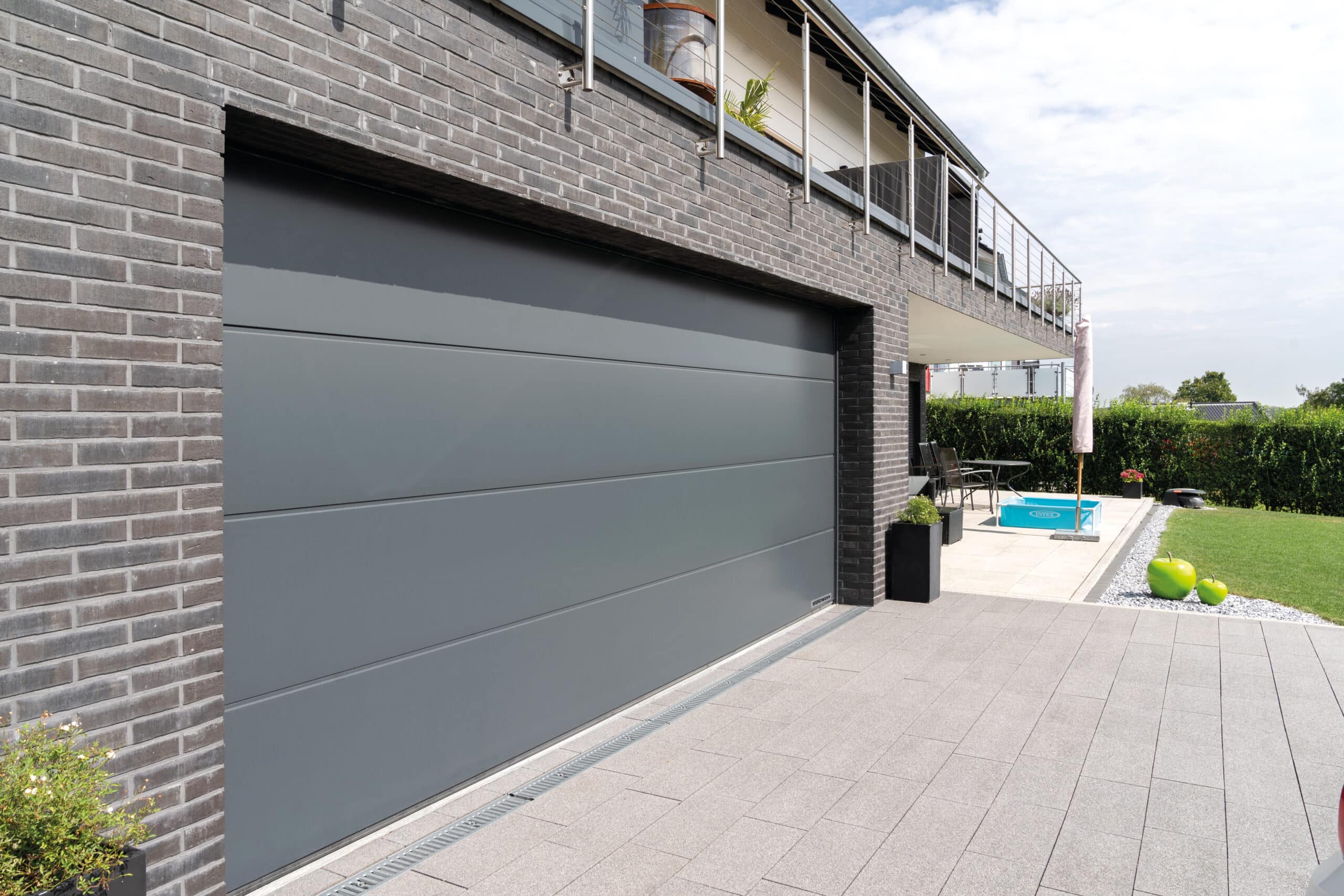 Modernes graues Garagentor an einem Backsteinhaus mit Terrasse, Pool und Garten im Hintergrund.