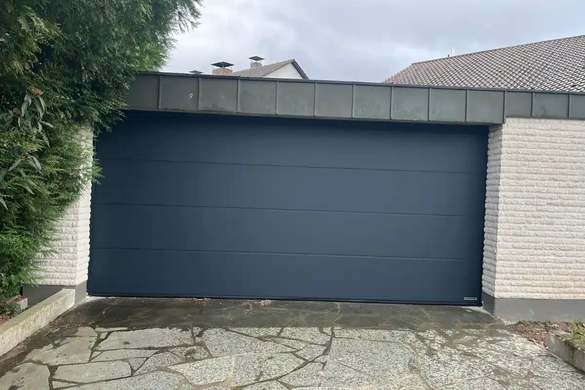 Ein geschlossenes, modernes dunkelgraues Garagentor in einer hellen Backsteinmauer mit einer Steinauffahrt davor.