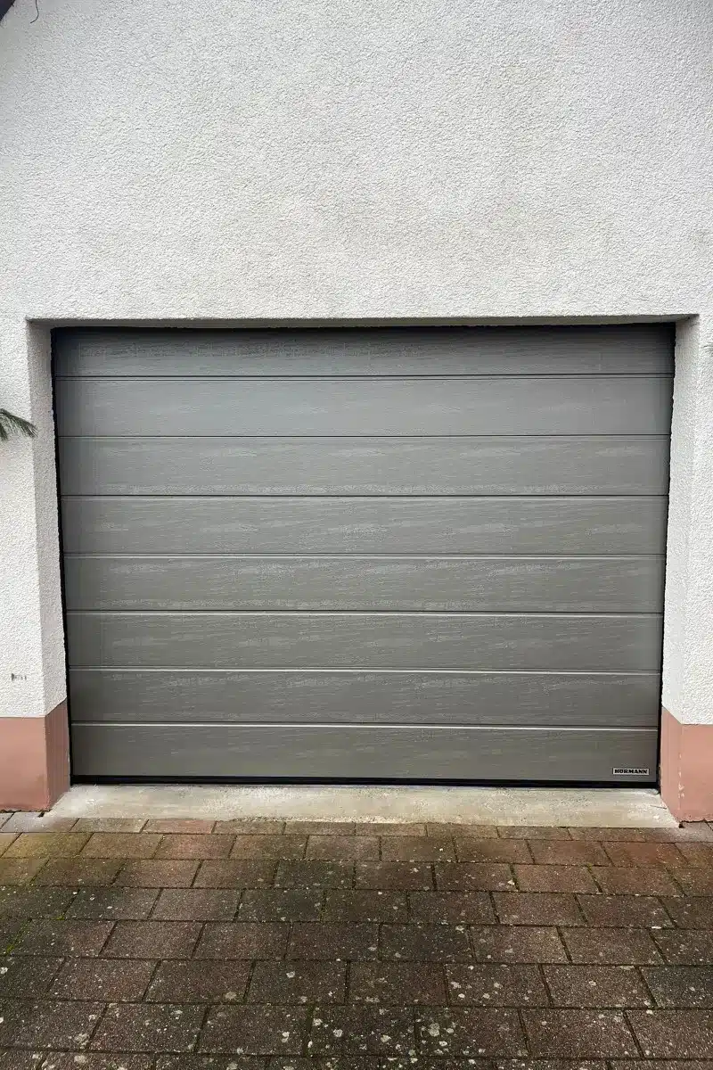 Geschlossenes graues Garagentor in einer weißen Außenwand, davor eine gepflasterte Einfahrt.