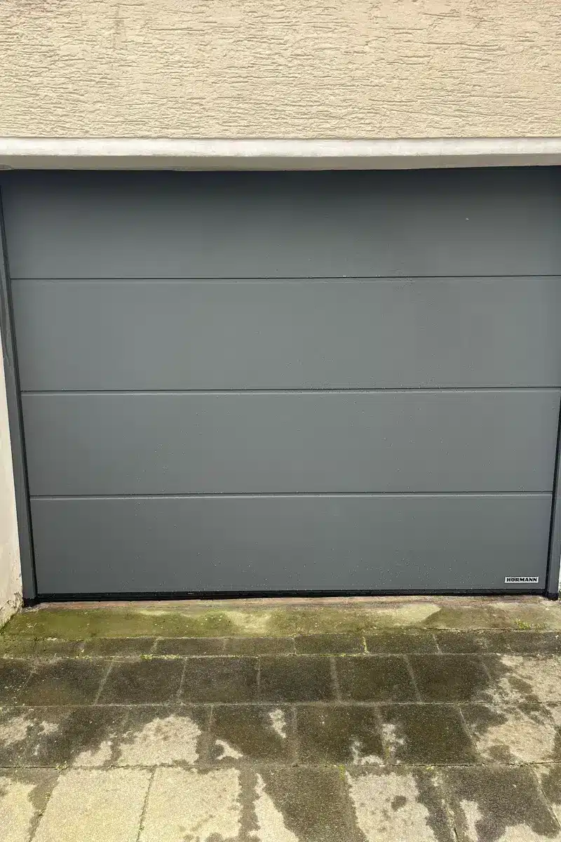 Ein geschlossenes graues Garagentor mit horizontalen Paneelen über einem moosbewachsenen, verwitterten Pflaster.