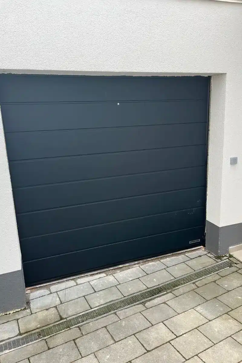 Ein schräg eingebautes Garagentor neben einer hellen Wand und grauem Steinpflaster.