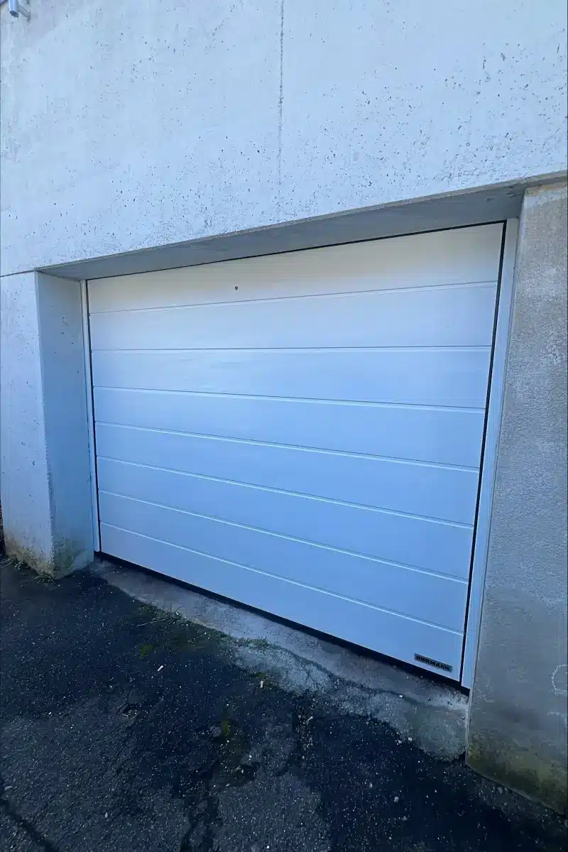 Ein geschlossenes weißes Garagentor in einer Betonmauer mit einer gepflasterten Auffahrt davor.