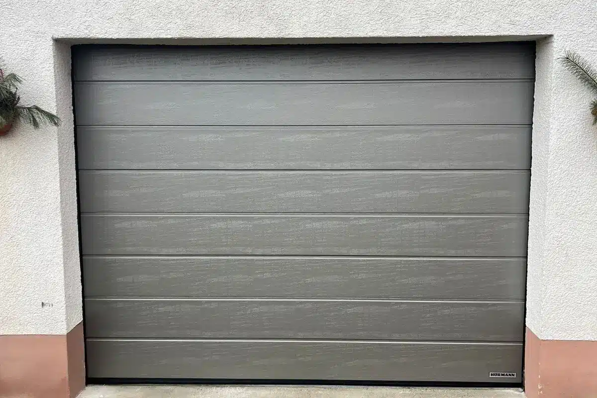 Geschlossenes graues Garagentor mit horizontalen Paneelen, die in eine weiße und rosafarbene Wand eingelassen sind.