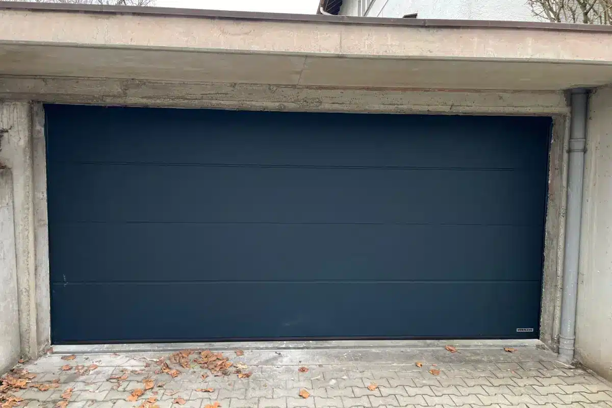 Geschlossenes modernes dunkelblaues Garagentor mit Betonrahmen und Laub auf dem Boden davor.