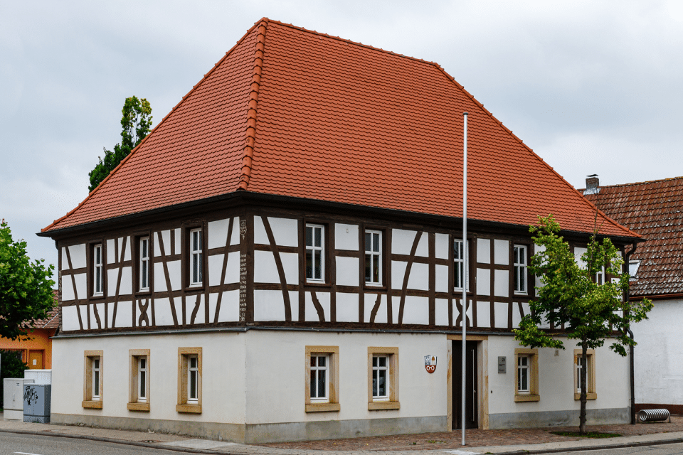 Traditionelles Fachwerkhaus mit rotem Ziegeldach und beigen Wänden an einer Straßenecke.